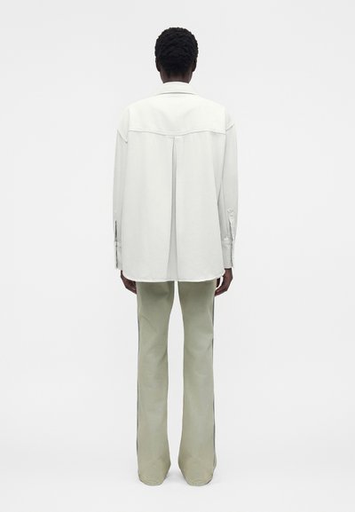 Άτομο με κοντά σγουρά μαλλιά που φοράει ένα άνετο λευκό πουκάμισο με μακριά μανίκια και ανοιχτό μπεζ φαρδιά παντελόνια, στέκεται γυρισμένο προς τα πίσω σε απλό φόντο.