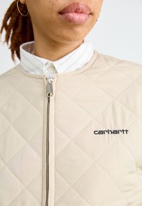Giacca trapuntata beige con chiusura frontale con zip, indossata su una camicia bianca con colletto e un logo nero ricamato.