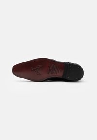 Suela de zapato de cuero negro con un acabado suave y brillante. Presenta un lado inferior marrón rojizo, un grabado decorativo y pequeñas tachas metálicas.