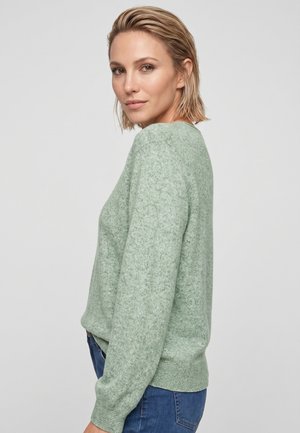 Frau mit kurzen blonden Haaren, die einen hellgrünen Pullover und blaue Jeans trägt, leicht seitlich gedreht vor einem schlichten Hintergrund.
