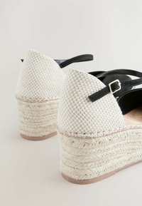 Sandale cu toc înalt, cu un brâu din piele neagră, partea superioară țesută în alb și o talpă din iută naturală. Texturate, cu un design elegant.