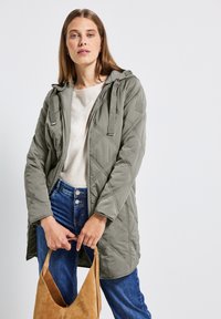 Veste matelassée olive avec capuche, présentant des coutures diagonales, une fermeture éclair à l'avant et deux poches verticales. Associée à un jean en denim et un sac beige.