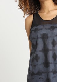 Kvinna med lockigt hår som bär en ärmlös svart och grått tie-dye klänning med genomskinlig svart överdel mot en enkel vit bakgrund.