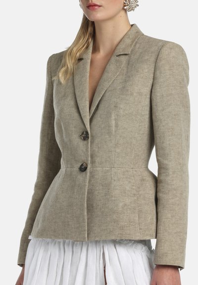 Mujer con blazer de lino beige entallado de dos botones, combinado con una falda blanca plisada y grandes pendientes decorativos.