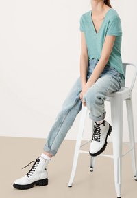 Mintgrünes V-Ausschnitt-T-Shirt, hellblaue schmal zulaufende Jeans und weiße Schnürstiefel mit schwarzen Sohlen, die auf einem Metallhocker sitzen.