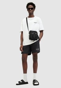 Μαύρο σορτσάκι με λογότυπο "ALLSAINTS", oversized λευκή μπλούζα, μαύρη τσάντα χιαστί και μαύρα σανδάλια slide με άσπρες λεπτομέρειες.