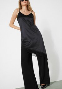 Vestido de satén negro con bajo asimétrico con borde de encaje, combinado con pantalones negros sueltos y zapatos negros de tacón.