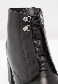 Bottine en cuir noir à talon haut, bout rond, avec fermeture à lacets maintenue par des œillets et crochets en métal argenté.