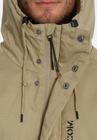 Beige gewatteerde jas met metalen drukknoopsluitingen, ritssluiting en trekkoord. Heeft een gladde textuur en opvallende stiksels aan de voorkant.