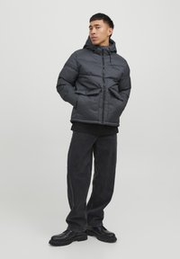 Sort polstret jakke med hætte, med en frontlynlås, to sidelommer og en mat finish. Båret over en sort skjorte og mørke jeans.