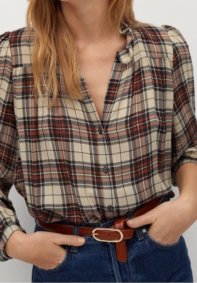 Femme portant une chemise à boutons à carreaux marron, beige et noir, rentrée dans un jean bleu avec une ceinture en cuir marron, les mains dans les poches.