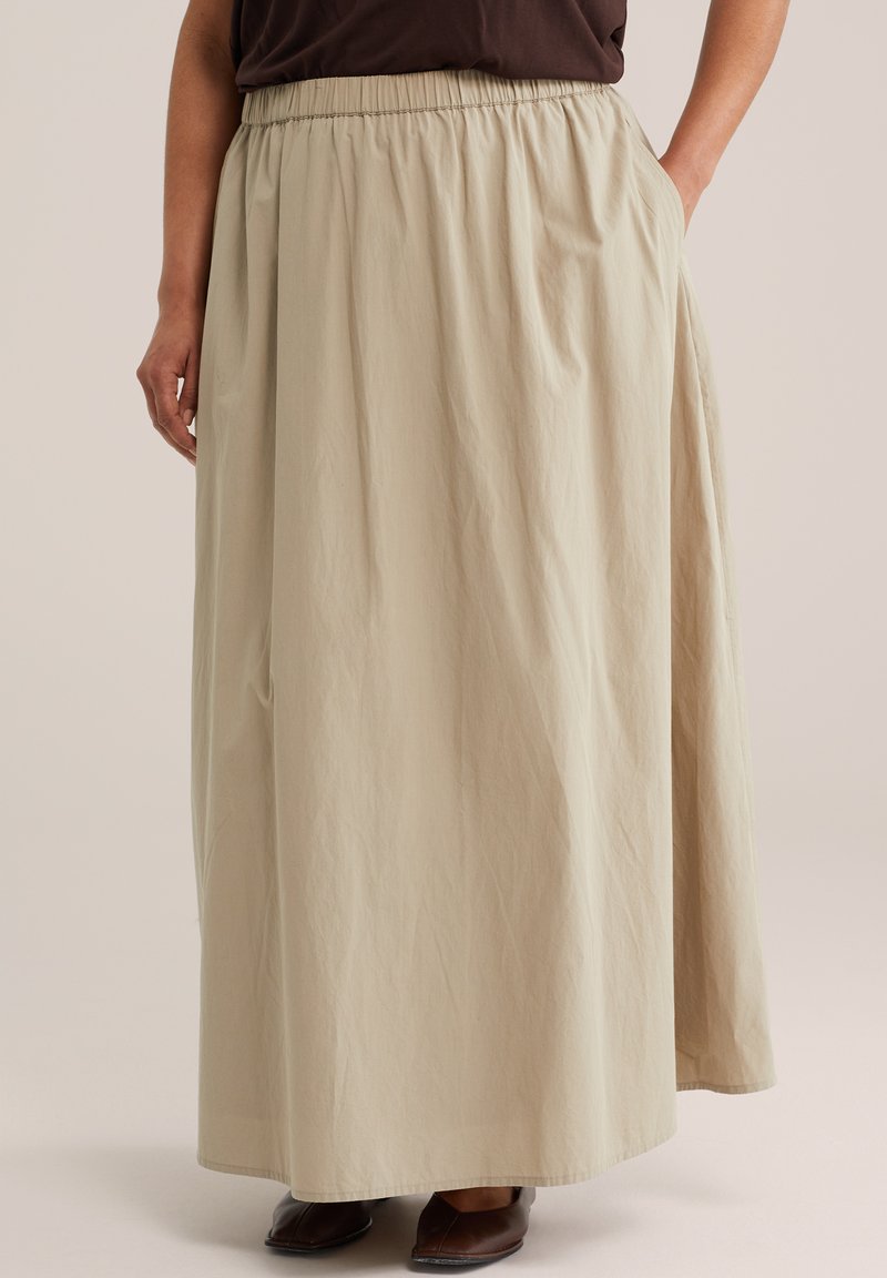 Femme portant une jupe beige, longue et ample, avec une taille élastique, assortie à des chaussures marron et un haut marron foncé rentré dedans.