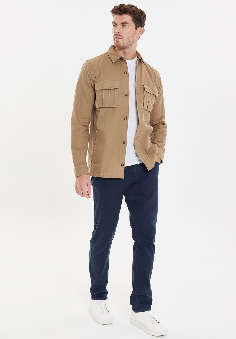 Hellbraune Baumwollhemdjacke mit zwei Brusttaschen, kombiniert mit navyblauen Hosen und weißen Sneakers, vor einem schlichten Hintergrund.