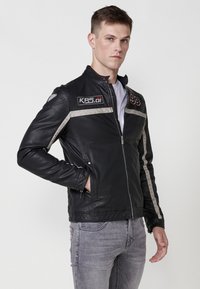Čierna kožená motocyklistická bunda so striebornými pruhmi, so zipsom, so šnúrkami na pleciach a detailnými logami. Nosená cez bielu košeľu a šedé džínsy.