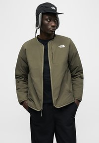 Νεαρός άντρας που φοράει μπουφάν χακί με φερμουάρ και μαύρο καπέλο τύπου «trapper» με αυτιά, τα χέρια στις τσέπες, κοιτάζοντας πλάγια.