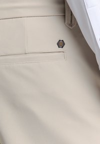 Beige broek van gladde stof, voorzien van een achterzak met een zwart en metaalachtige hexagonaal accent en een streeppatroon.