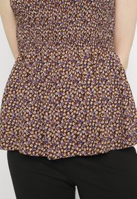 Květinový peplum top v béžové barvě, s nařaseným korzetem a fialovými a oranžovými květinami. Nosený přes černé kalhoty, s rozšířeným lemem.