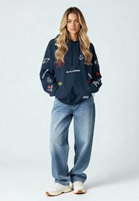 Felpa con cappuccio blu navy con vari ricami colorati, abbinata a jeans baggy azzurri e scarpe da ginnastica bianche.