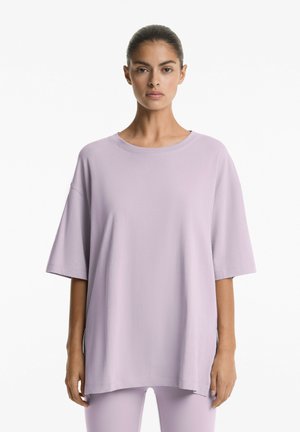 Tricou oversized de culoare lavandă deschisă, fabricat dintr-un material moale, cu guler rotund și umeri lăsați, cu mâneci scurte și slituri pe laterale.