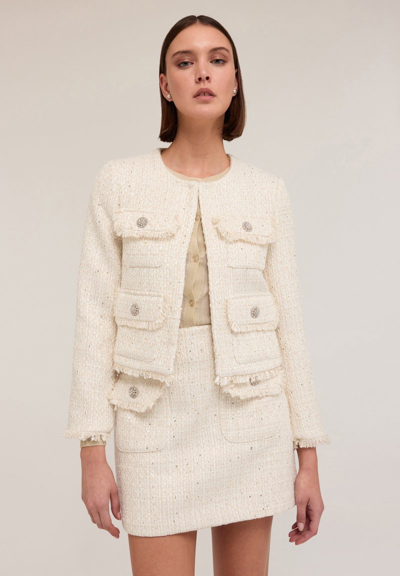 Veste en tweed blanc texturé avec poches avant et bords frangés, assortie d'une jupe correspondante. Les deux pièces sont ornées de boutons décoratifs.