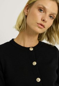 Cardigan noir avec un col rond, orné de cinq boutons décoratifs dorés sur le devant. Tissu texturé, design ajusté.