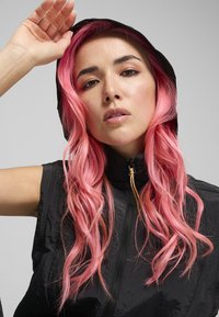 Gilet à capuche sans manches noir avec un col haut, doté d'une fermeture éclair sur le devant et d'une texture brillante, porté par une personne aux longs cheveux roses.