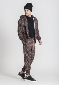 Chándal marrón con un patrón y acentos de logo, que incluye una chaqueta con cremallera y pantalones con cintura elástica, acompañado de un gorro negro y zapatillas.