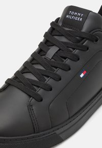 Černé kožené tenisky s plochými černými tkaničkami, na jazyku s bílým logem "TOMMY HILFIGER" a malým červeným, bílým a modrým emblémem.