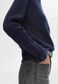 Navyblå stickad tröja med en strukturerad yta, med hög krage och ribbade ärmslut, tillsammans med mörkgrå denimjeans.