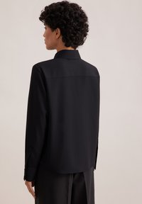 Camicia nera a maniche lunghe con collo, caratterizzata da una texture liscia e cucitura sul giogo posteriore. Polsini con bottoni che aggiungono dettagli al design minimalista.