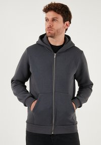 Sivi dukserica na zip s mekim materijalom, s prednjim zatvaračem, dva džepa i rebrastim donjim rubom. Model s kapuljačom i dugim rukavima.