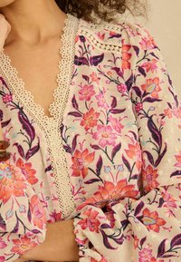 Kvetinová blúzka z ľahkej látky so vzorom ružových a oranžových kvetov, výstrih do V s čipkovým lemom a volánovými rukávmi.
