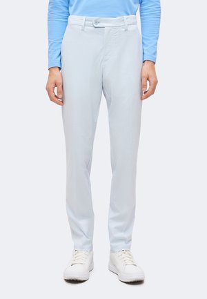 Homme portant un pantalon ajusté bleu clair, des baskets blanches et une chemise bleu clair à manches longues, debout devant un fond uni.