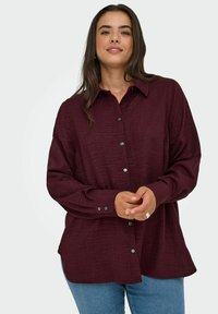 Burgundská košile na knoflíky s texturovanou tkaninou, límečkem a dlouhými rukávy, spárovaná s světle modrými džíny. Má černé knoflíky.