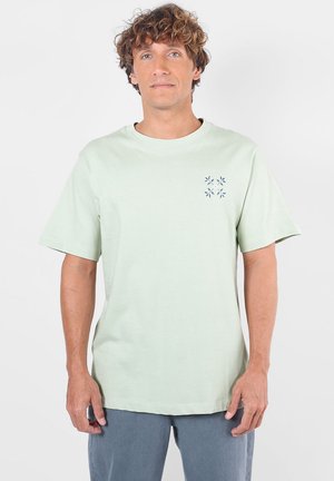 Homme aux cheveux bouclés portant un t-shirt vert clair à manches courtes avec un petit motif floral bleu sur la poitrine gauche, et un pantalon gris.