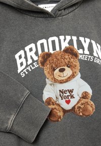 Felpa grigia con un grafico di un orso di peluche in camicia bianca con la scritta "New York" e un cuore. Include dettagli con polsini a coste e lettering audace.