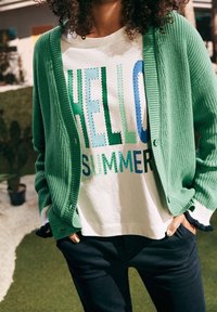 Osoba v zeleném kardiganu přes bílé tričko s barevným nápisem "AHOJ LÉTO", stojící venku s rukama v kapsách tmavých kalhot.