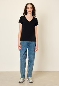 Mladá žena s kudrnatými vlasy, oblečená v černém tričku s výstřihem do V, volných modrých džínách a stříbrných teniskách, stojí na světlé podlaze před jednobarevnou zdí.