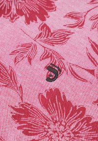 Textiel met een roze achtergrond en een geweven bloemenpatroon in het rood. Bevat een klein zwart logo dat dicht bij het midden is geplaatst.