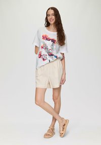 Witte T-shirt met een kleurrafloraal grafisch ontwerp en tekst, gecombineerd met lichtbeige, gestreepte shorts en tan kleurige sandalen met bandjes. Minimalistisch ontwerp.