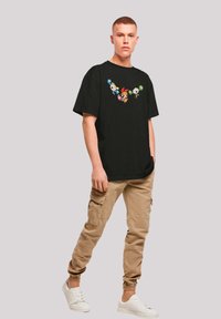 Schwarzes Baumwoll-T-Shirt mit einem bunten Cartoon-Charakterdesign auf der Brust, kombiniert mit beigen Cargo-Hosen und weißen Sneakers.