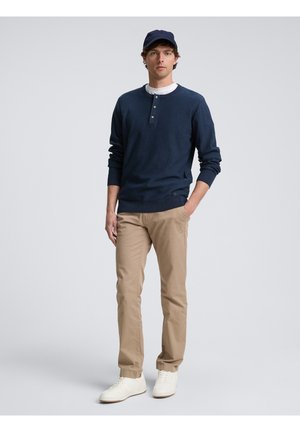 Mornarski henley pulover s tri gumba, bež chinos hlače i bijele tenisice; model nosi mornarsku kapu; glatka tkanina, ležeran kroj.