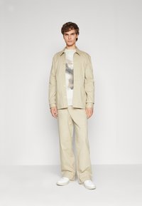 Chaqueta beige clara sobre un suéter blanco de estampado, combinada con pantalones beige y zapatillas blancas. Material suave con un ajuste relajado.