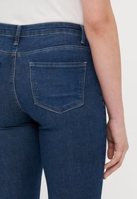 Tamno plave traperice s uskim krojem, s visokim pojasom, dva stražnja džepa i kontrastnim šavovima.