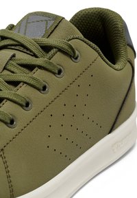 Nahaufnahme eines olivgrünen Sneakers mit strukturiertem Obermaterial, passenden Schnürsenkeln, durchlöcherten Seitenverzierungen, gepolstertem Kragen und weißer Sohle.