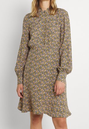 Femme portant une robe fleurie à manches longues, longueur genoux, avec un col montant et des poignets boutonnés sur fond uni.