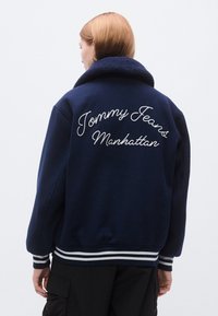 Tamsiai mėlynas flisinis bombonešio stiliaus švarkas su dideliu išsiuvinėtu užrašu „Tommy Jeans Manhattan“ ant nugaros; turi šerpą apykaklę ir dryžuotas rankogalius.