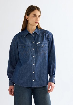 Donna che indossa una camicia di jeans blu con bottoni automatici e due tasche frontali, abbinata a jeans blu, in piedi davanti a uno sfondo bianco semplice.