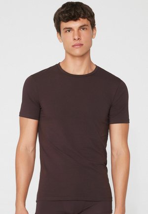 Junger Mann mit kurzen lockigen dunklen Haaren, der ein enganliegendes, einfarbiges dunkelbraunes Kurzarm-T-Shirt vor einem einfarbigen hellen Hintergrund trägt.