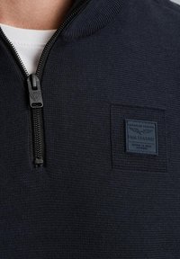 Felpa con zip navy in tessuto testurizzato, dotata di colletto alto, zip nera e un badge rettangolare sul petto.